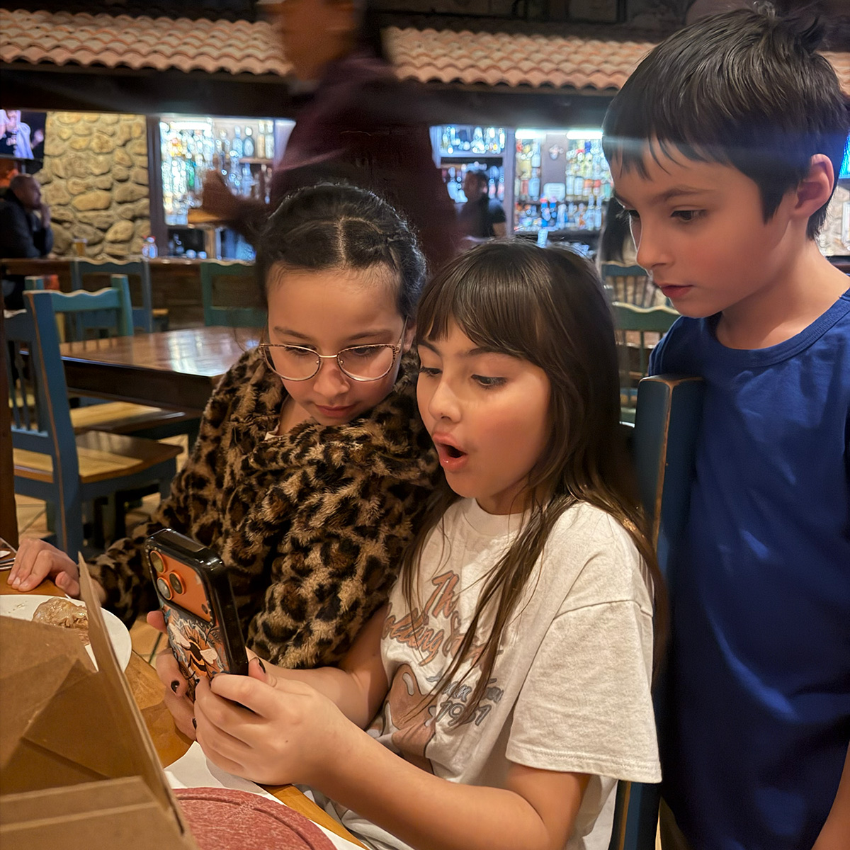 Three children excitedly looking at a phone in a restaurant.