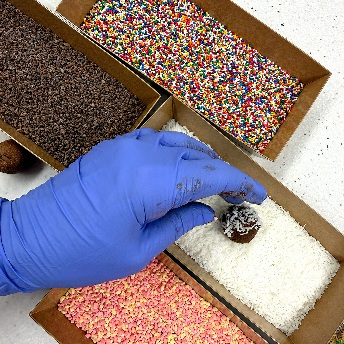 A gloved hand decorating a chocolate truffle with sprinkles.